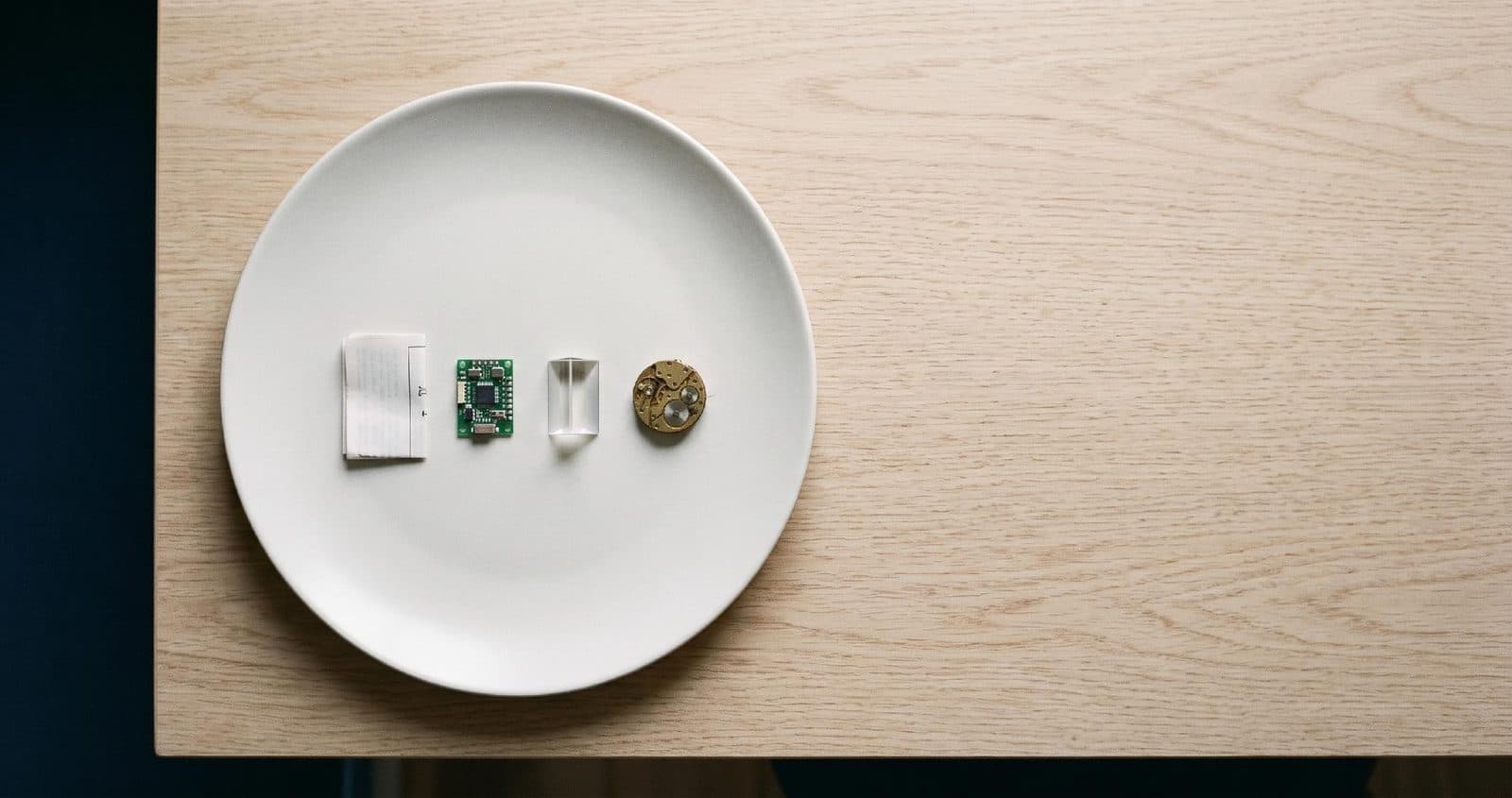 A plate holding four objects in a row: a folded paper form, a circuit board, a glass prism and a watch movement